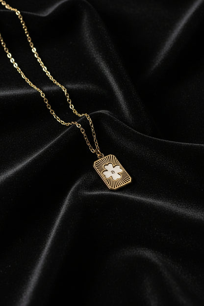 A gold-toned chain necklace with a rectangular pendant featuring a white clover design and gold sunburst details, displayed on a clean white background.